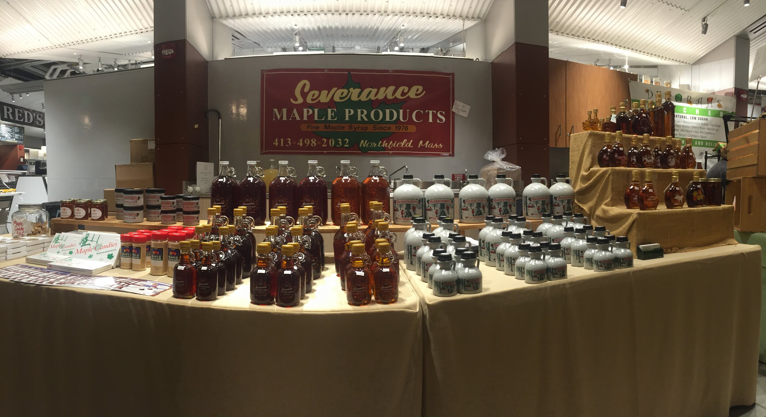 Display of Severance Maple Products featuring a variety of maple syrups and candies, showcasing bottles of different sizes and flavors, at a market in Northfield, Massachusetts. The banner prominently features the brand name and contact information.