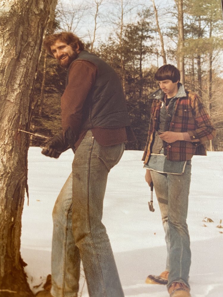 Two men working outdoors in winter, one using a tool on a tree and the other holding a hammer, surrounded by snow and forest. The men are dressed in casual, layered clothing typical of the 1970s, highlighting a rustic, outdoor lifestyle.