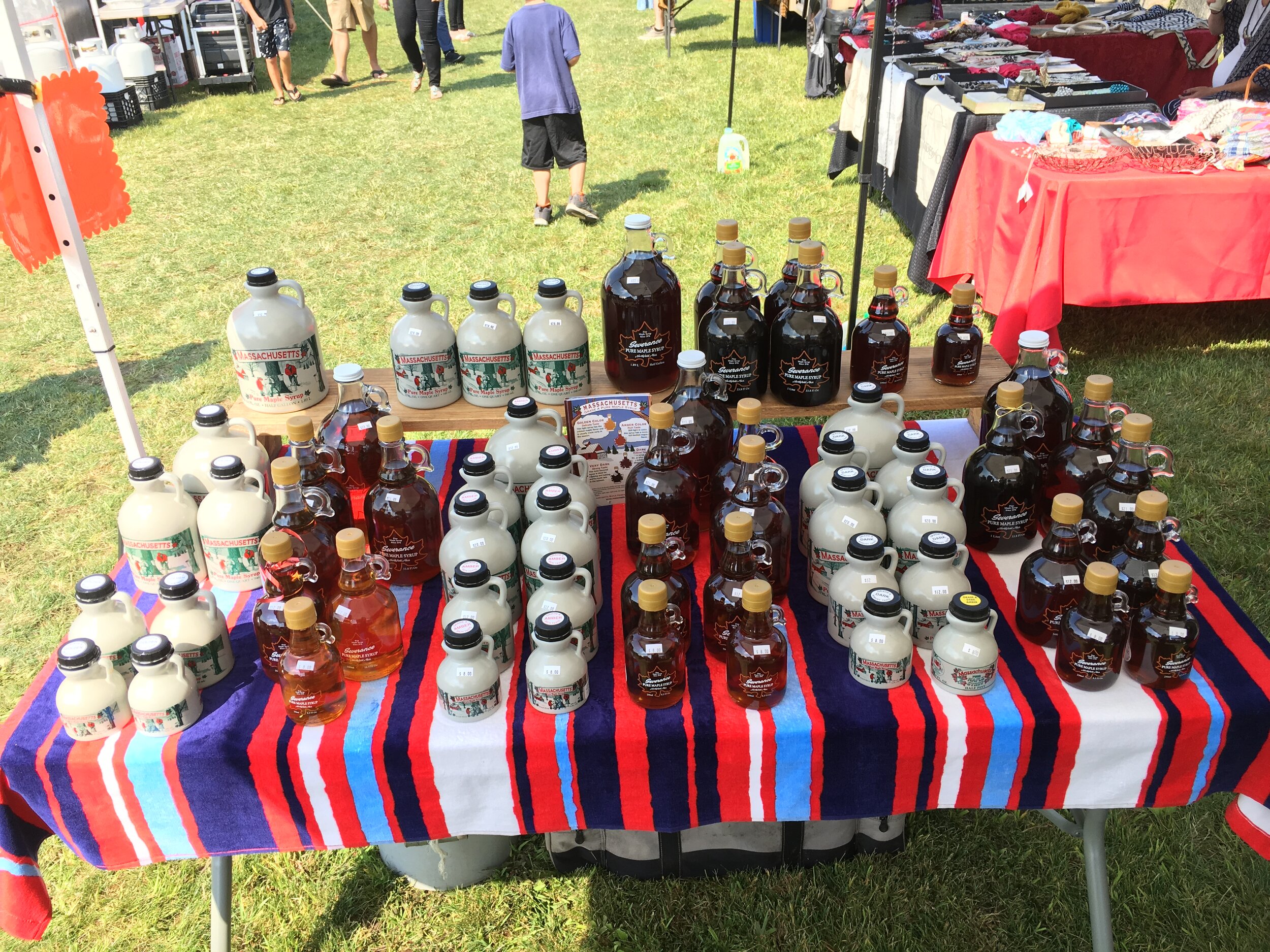 Display of various bottles of maple syrup on a colorful tablecloth at a local market, showcasing different grades and sizes of syrup. In the background, people are browsing nearby vendor stalls.