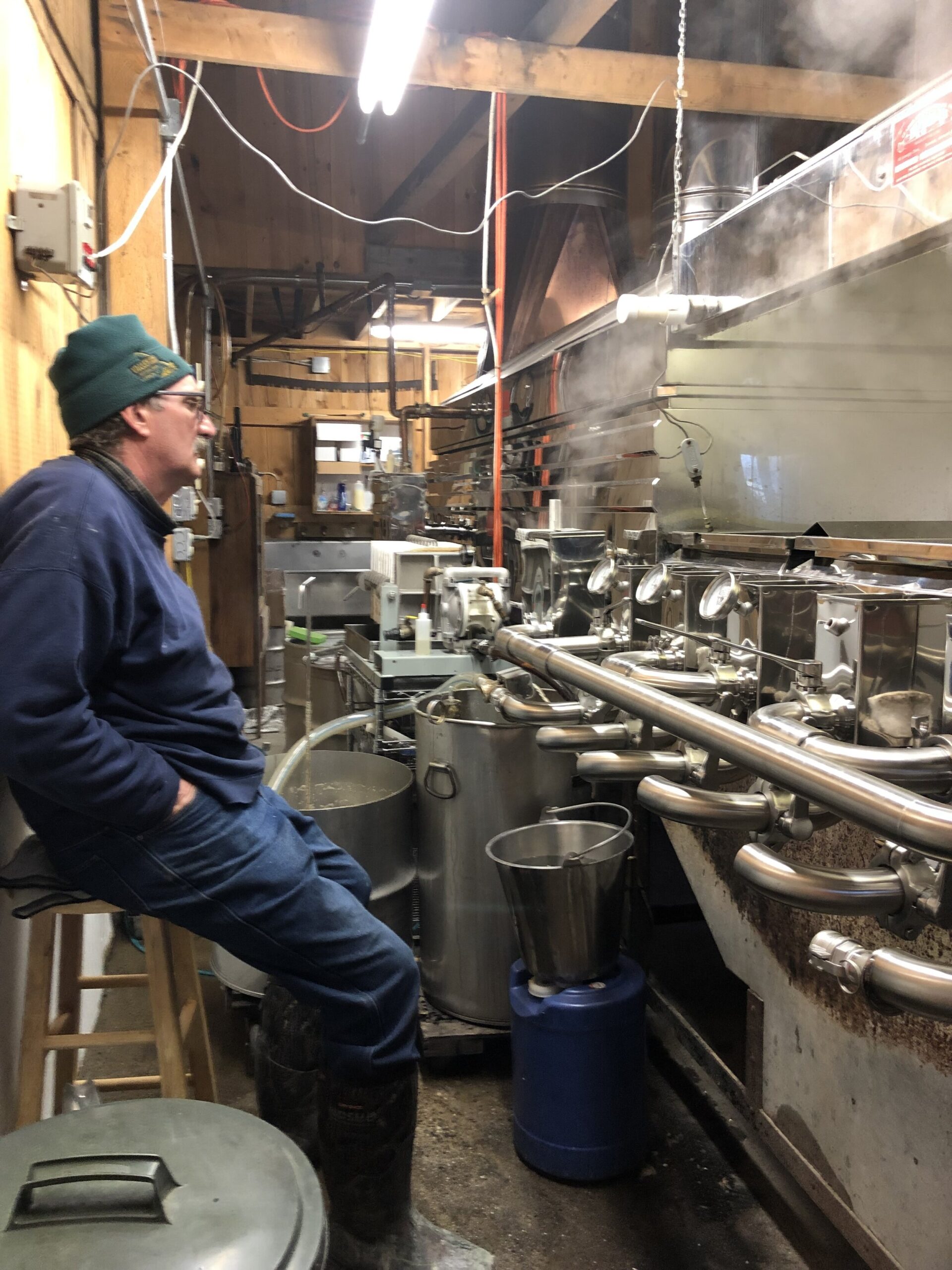 Man sitting on a stool in a sugar shack, observing the maple syrup production process with steam rising from the equipment. The interior features wooden beams and various stainless steel containers and machinery used for boiling sap.